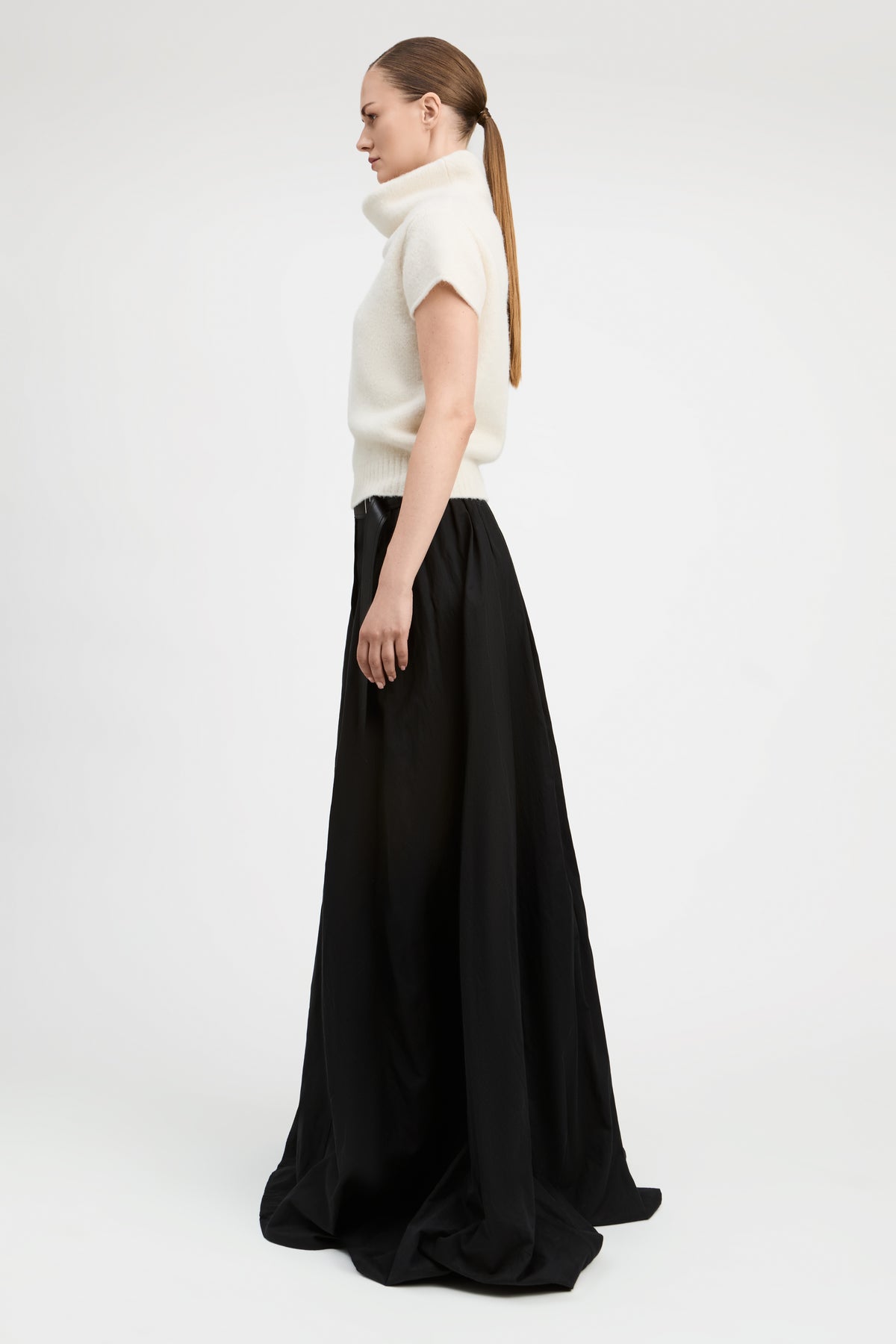 A woman in profile wearing a cream short-sleeve mock-neck knit top and a long black skirt, standing against a plain light gray studio background.