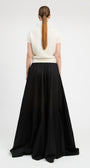A model shown from the back wearing a cream short-sleeve mock-neck knit top and a long black pleated skirt, photographed against a seamless white studio background.