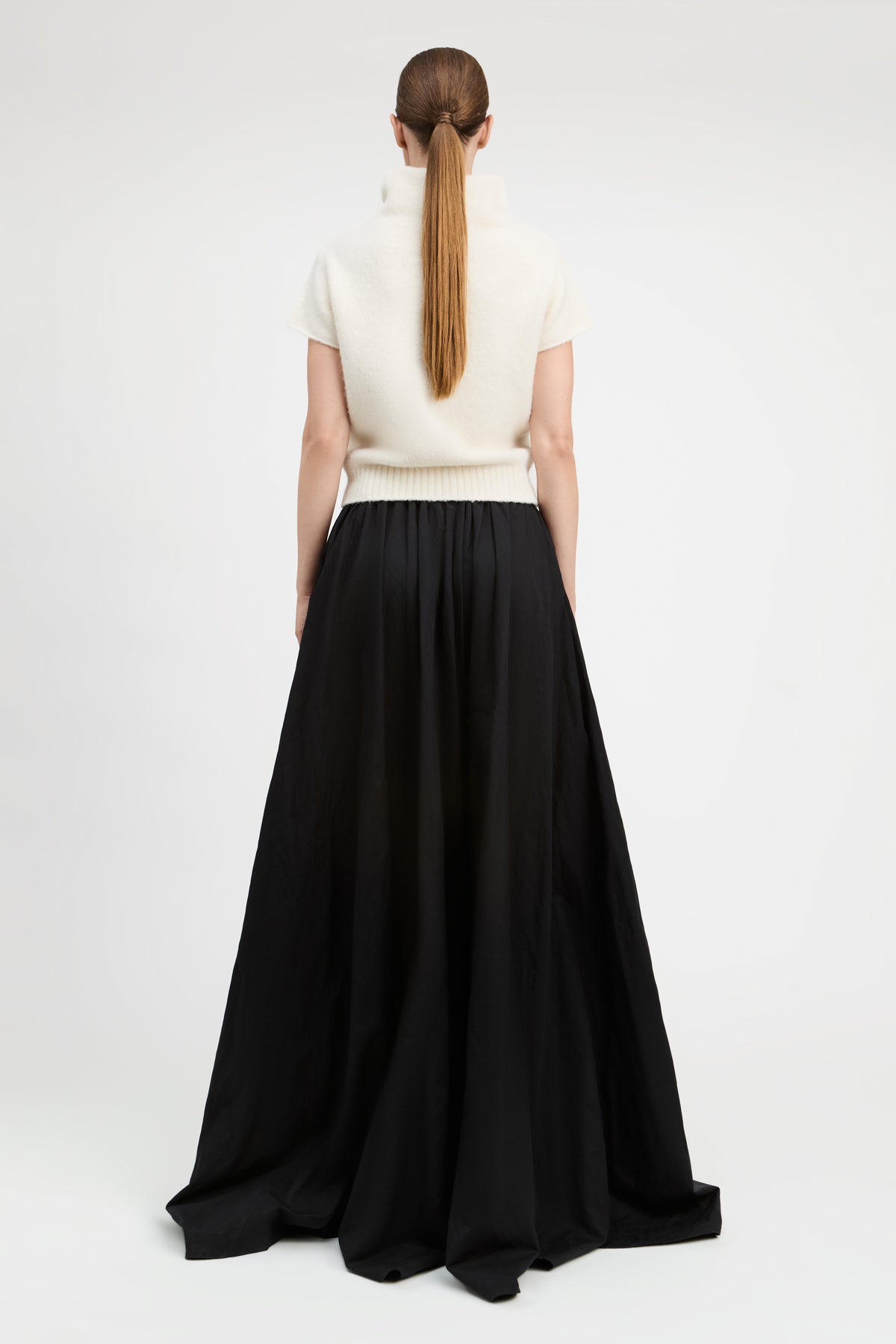 A model shown from the back wearing a cream short-sleeve mock-neck knit top and a long black pleated skirt, photographed against a seamless white studio background.