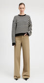 A model stands against a light gray studio background wearing a black and white horizontally striped knit sweater with black cuffs and hem, paired with wide-leg beige trousers.