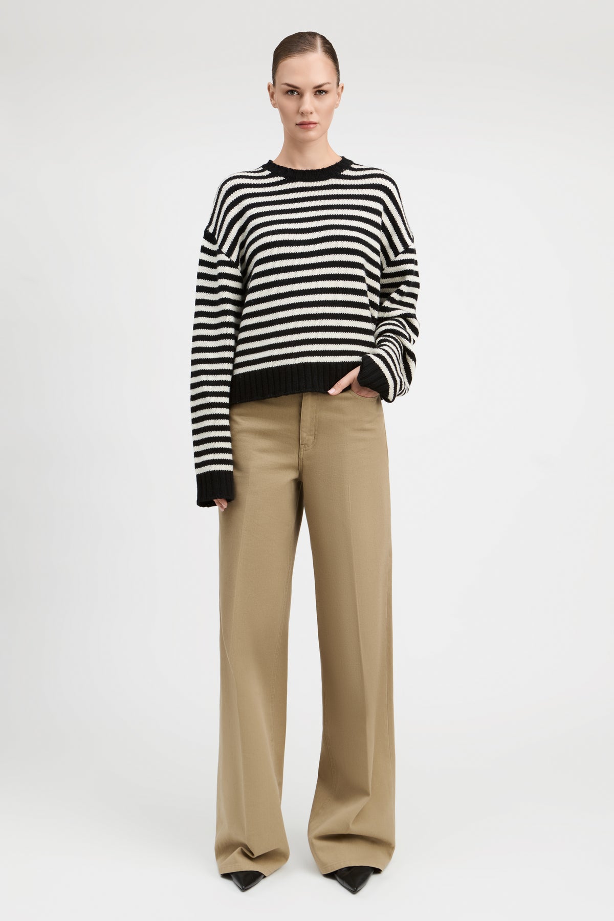 A model stands against a light gray studio background wearing a black and white horizontally striped knit sweater with black cuffs and hem, paired with wide-leg beige trousers.