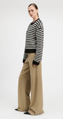 A woman modeling a black and white horizontally striped knit sweater with black cuffs and hem, paired with wide-leg beige trousers, against a plain light gray studio background.