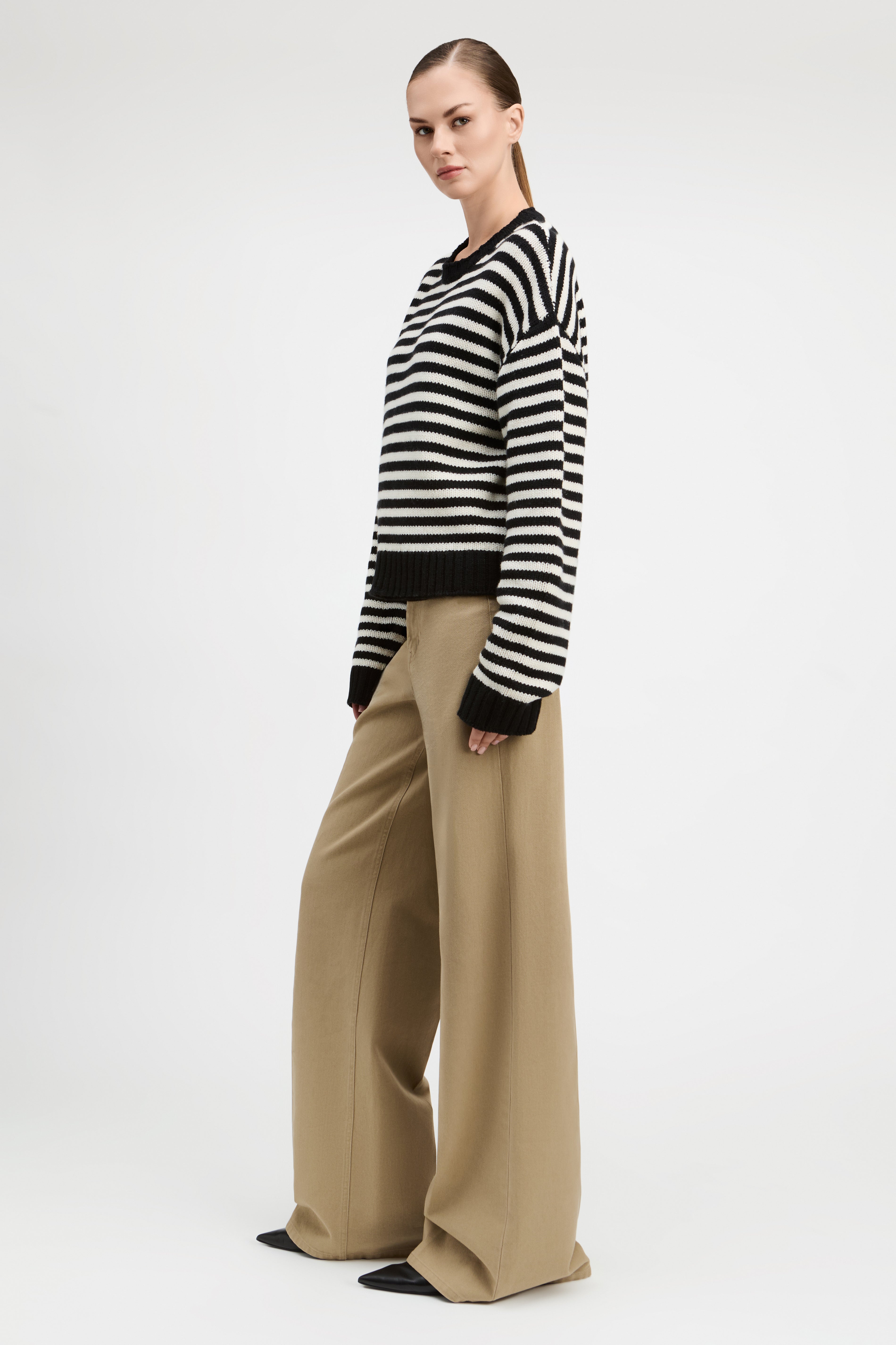 A woman modeling a black and white horizontally striped knit sweater with black cuffs and hem, paired with wide-leg beige trousers, against a plain light gray studio background.