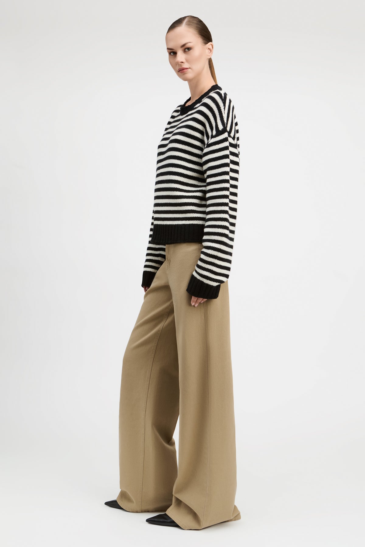 A woman modeling a black and white horizontally striped knit sweater with black cuffs and hem, paired with wide-leg beige trousers, against a plain light gray studio background.
