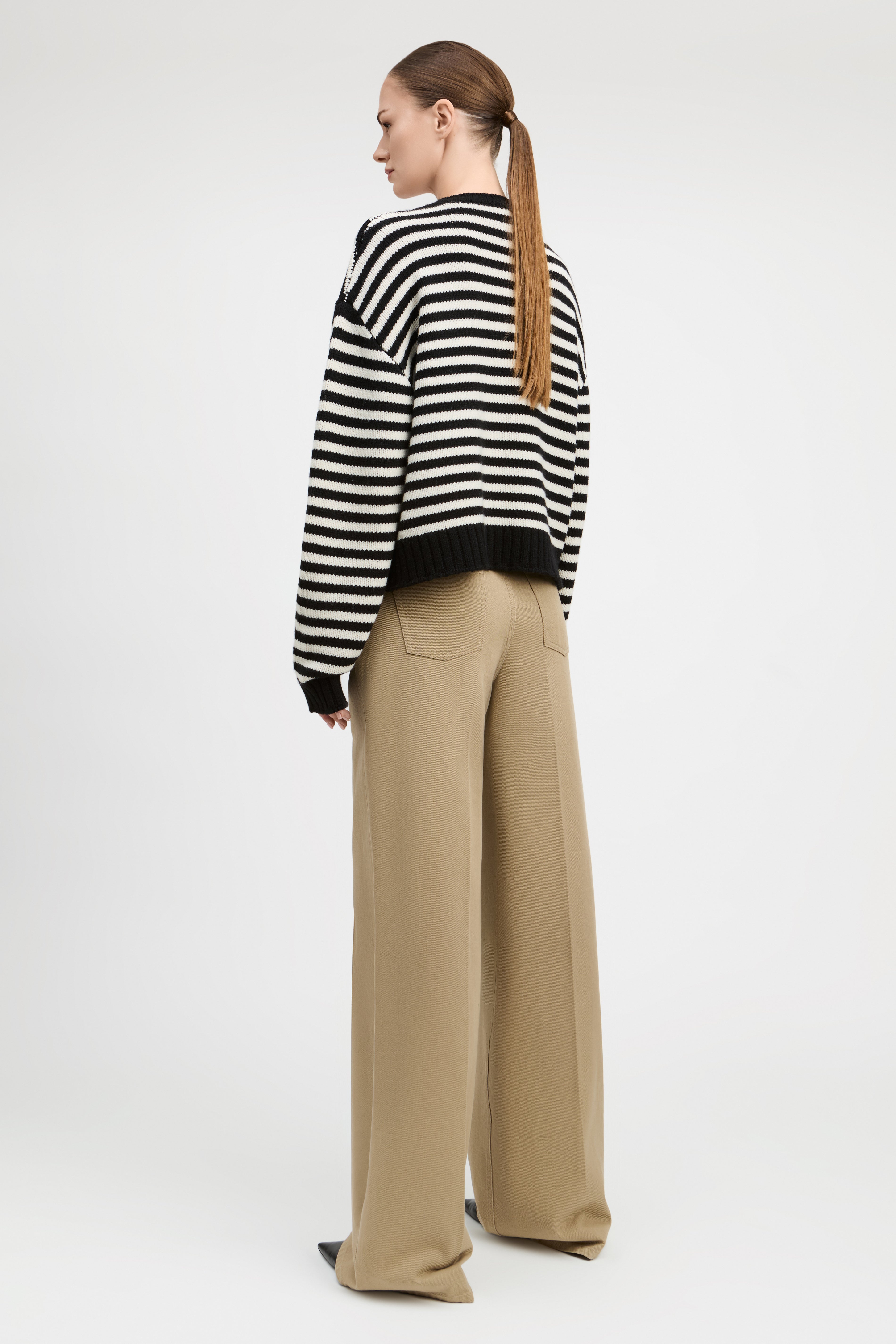 A woman shown from the back wearing a black and white striped knit sweater and beige wide-leg trousers, standing against a light gray studio background.
