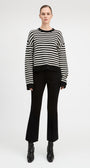 A model stands against a light gray studio background wearing a black and white horizontally striped knit sweater, black cropped flare pants, and black ankle boots.