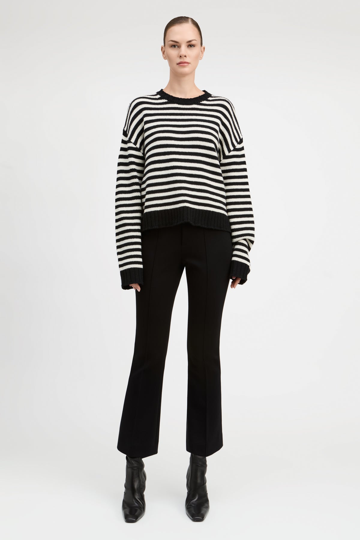 A model stands against a light gray studio background wearing a black and white horizontally striped knit sweater, black cropped flare pants, and black ankle boots.