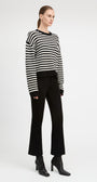 A model stands against a light gray studio background wearing a black and white horizontally striped long-sleeve knit sweater, black flared trousers, and black heeled ankle boots.