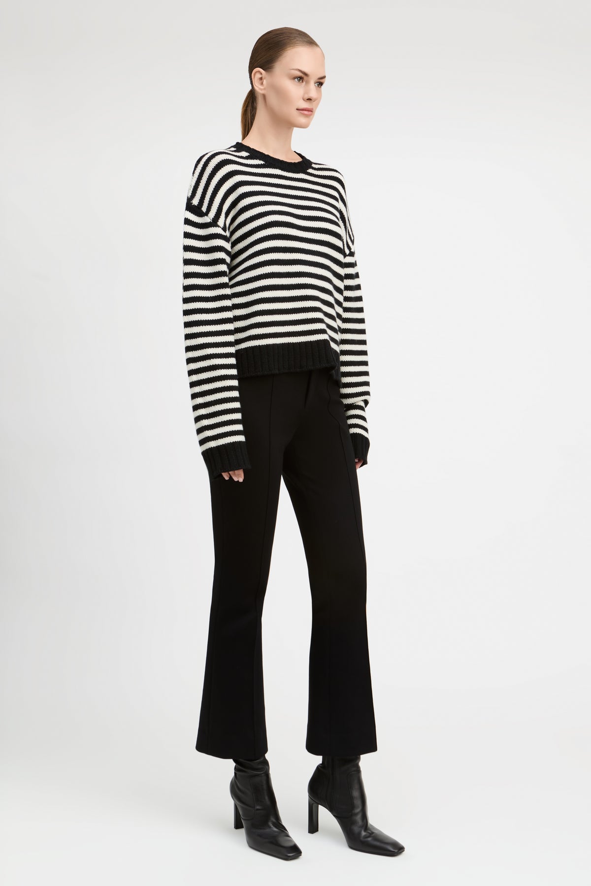 A model stands against a light gray studio background wearing a black and white horizontally striped long-sleeve knit sweater, black flared trousers, and black heeled ankle boots.