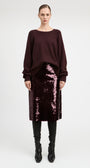 A model stands against a light gray studio background wearing a dark burgundy long-sleeve knit top, a matching sequined midi skirt, and black knee-high boots.