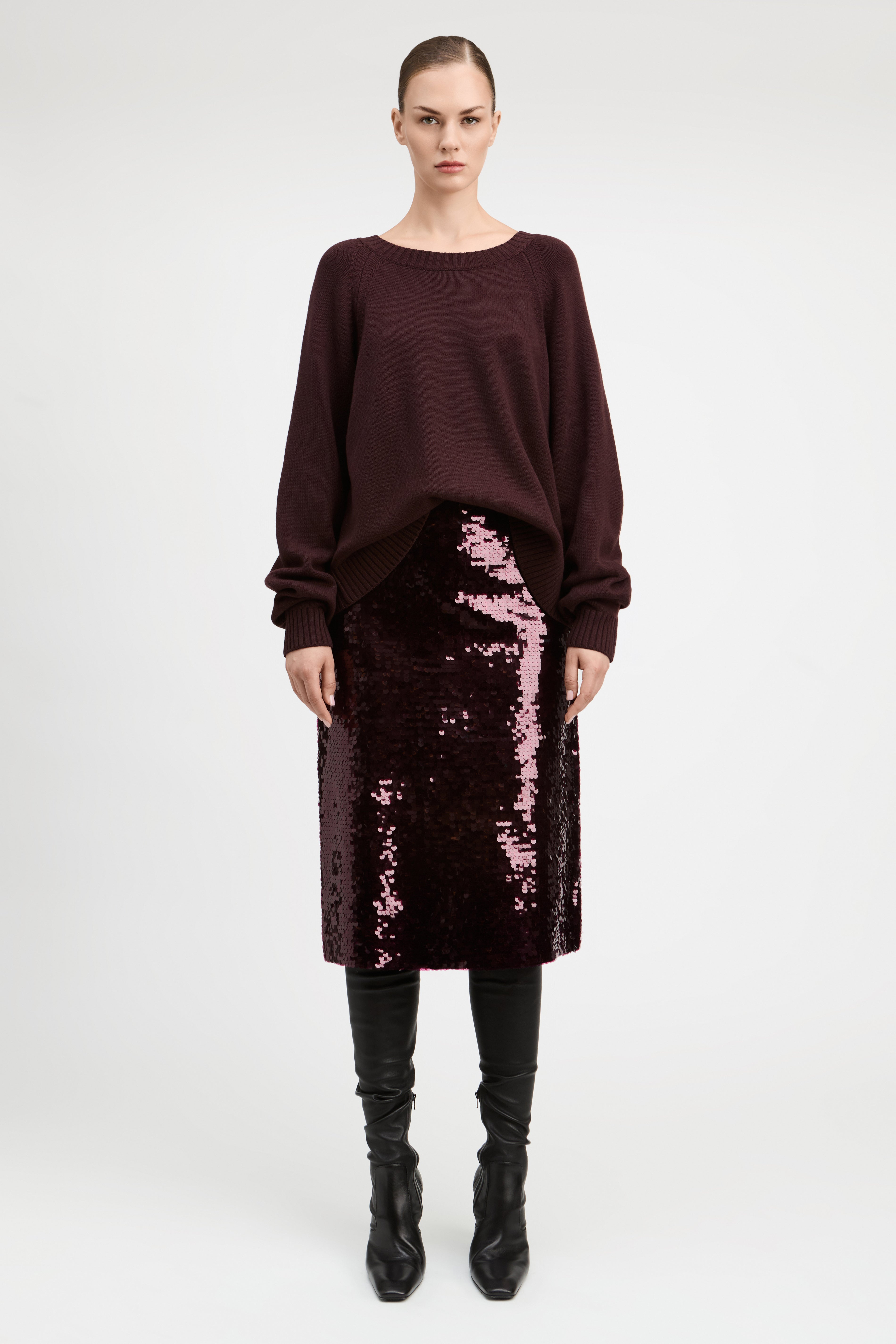 A model stands against a light gray studio background wearing a dark burgundy long-sleeve knit top, a matching sequined midi skirt, and black knee-high boots.