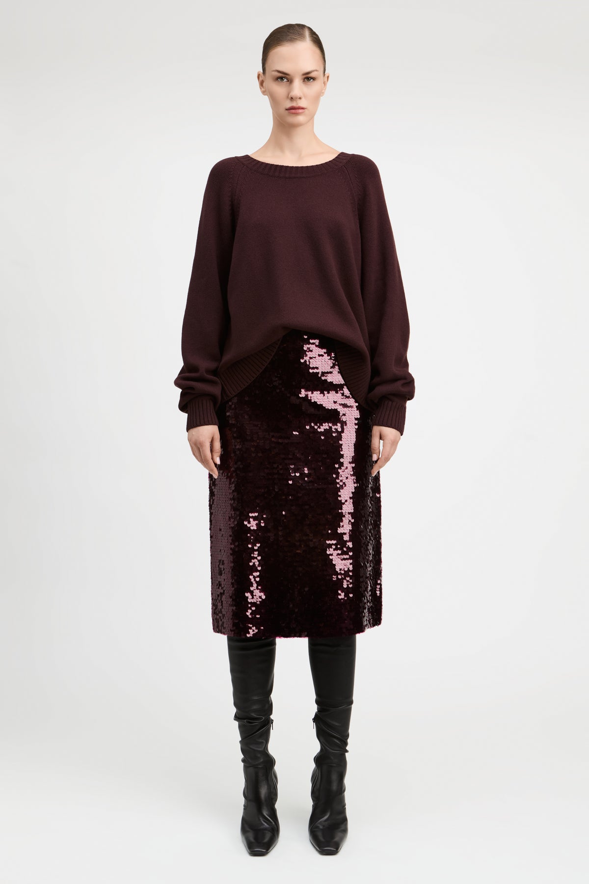 A model stands against a light gray studio background wearing a dark burgundy long-sleeve knit top, a matching sequined midi skirt, and black knee-high boots.