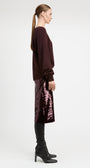 A woman in profile wearing a dark burgundy long-sleeve top, a sparkling mauve-pink sequined midi skirt, black leggings, and black heeled boots, photographed against a plain light gray studio background.
