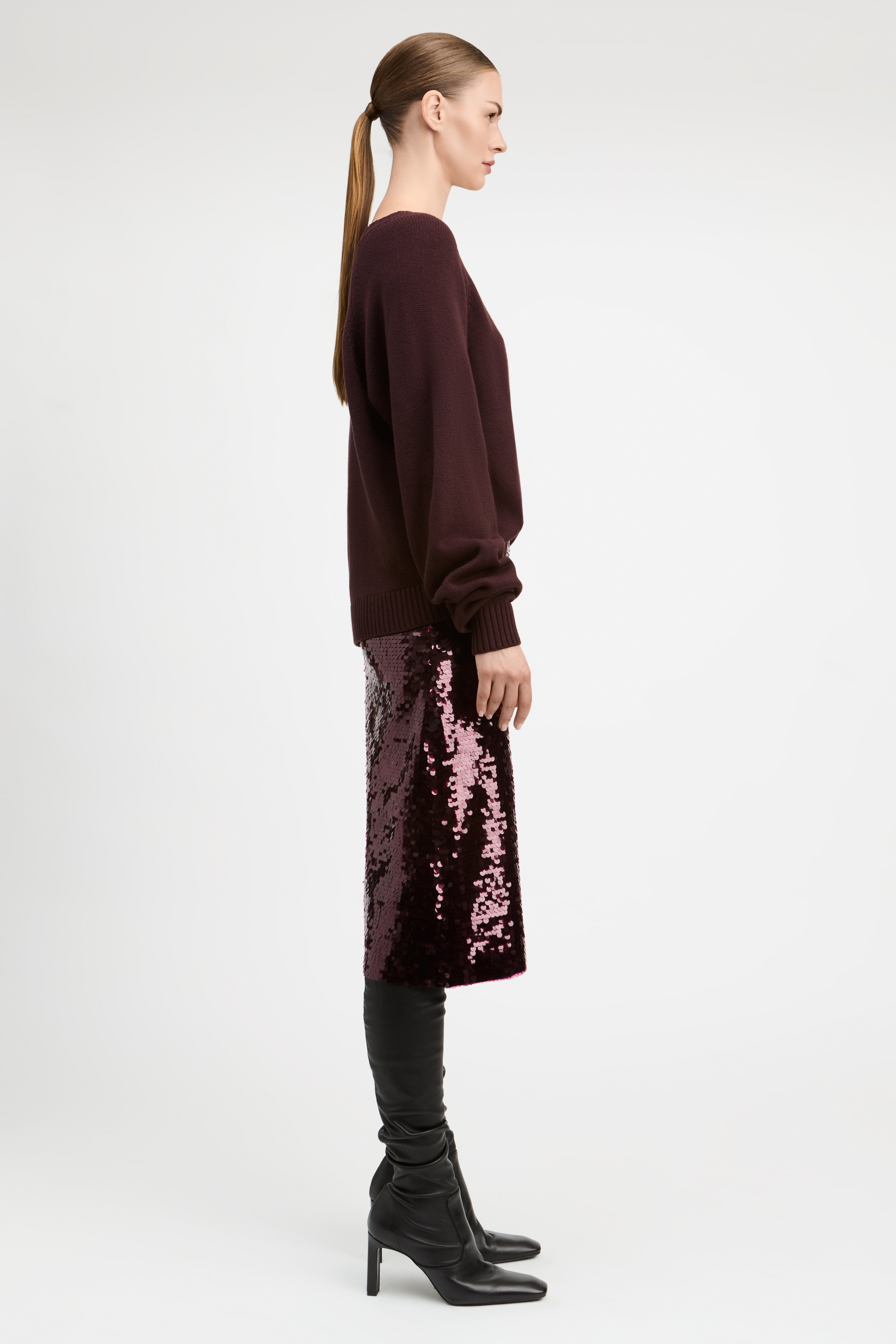A woman in profile wearing a dark burgundy long-sleeve top, a sparkling mauve-pink sequined midi skirt, black leggings, and black heeled boots, photographed against a plain light gray studio background.