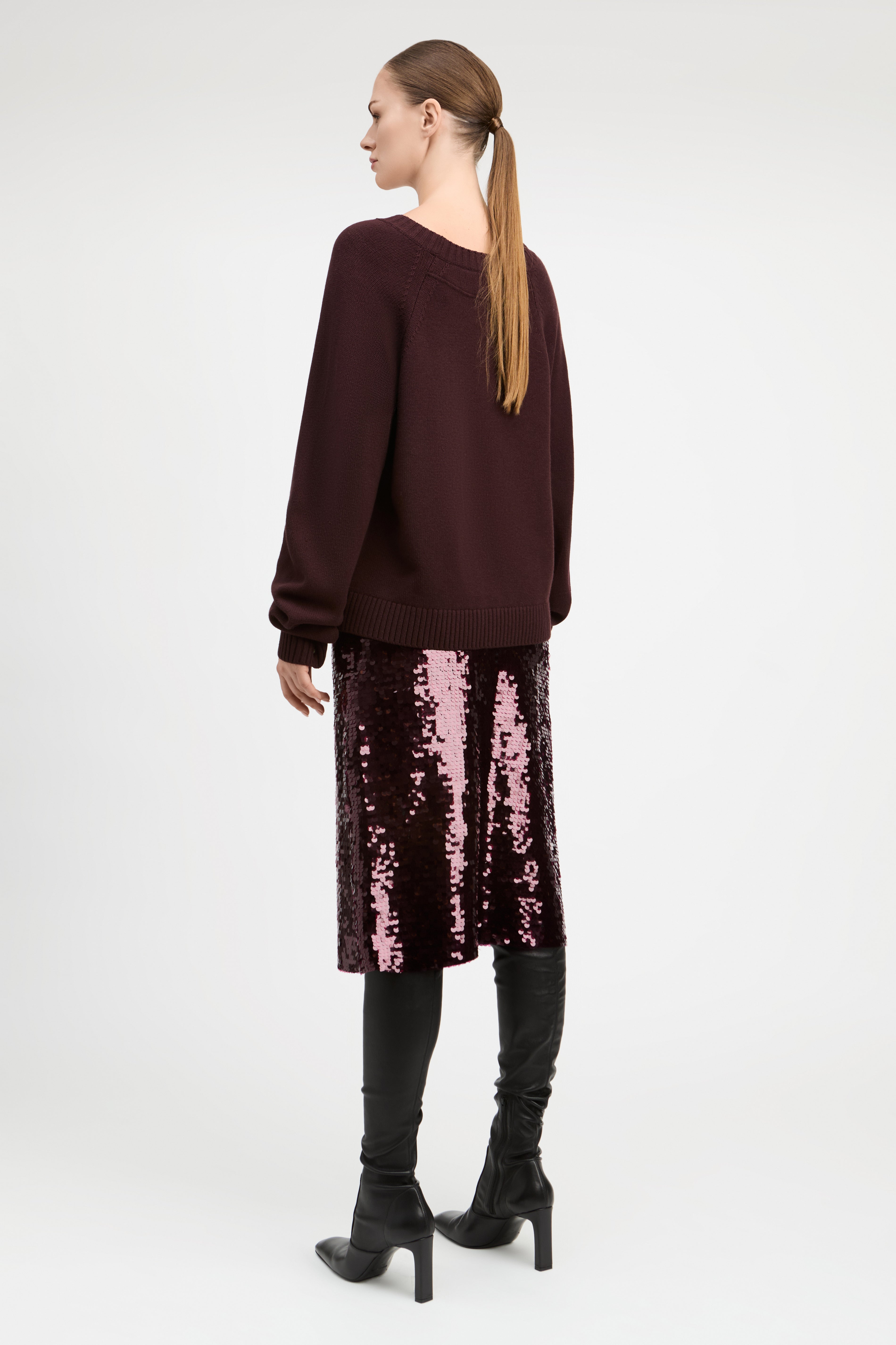 A woman shown from the back wearing a dark burgundy knit sweater, a pink sequined midi skirt, black over-the-knee boots, and black ankle boots, against a plain light gray studio background.