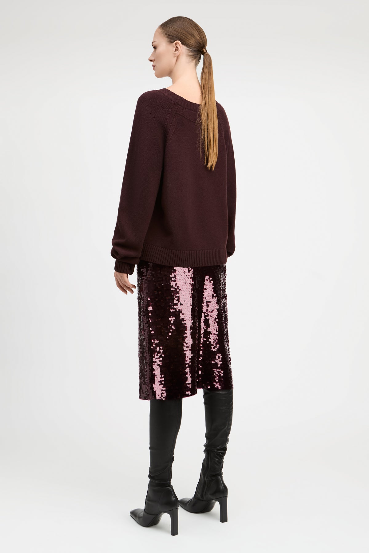 A woman shown from the back wearing a dark burgundy knit sweater, a pink sequined midi skirt, black over-the-knee boots, and black ankle boots, against a plain light gray studio background.