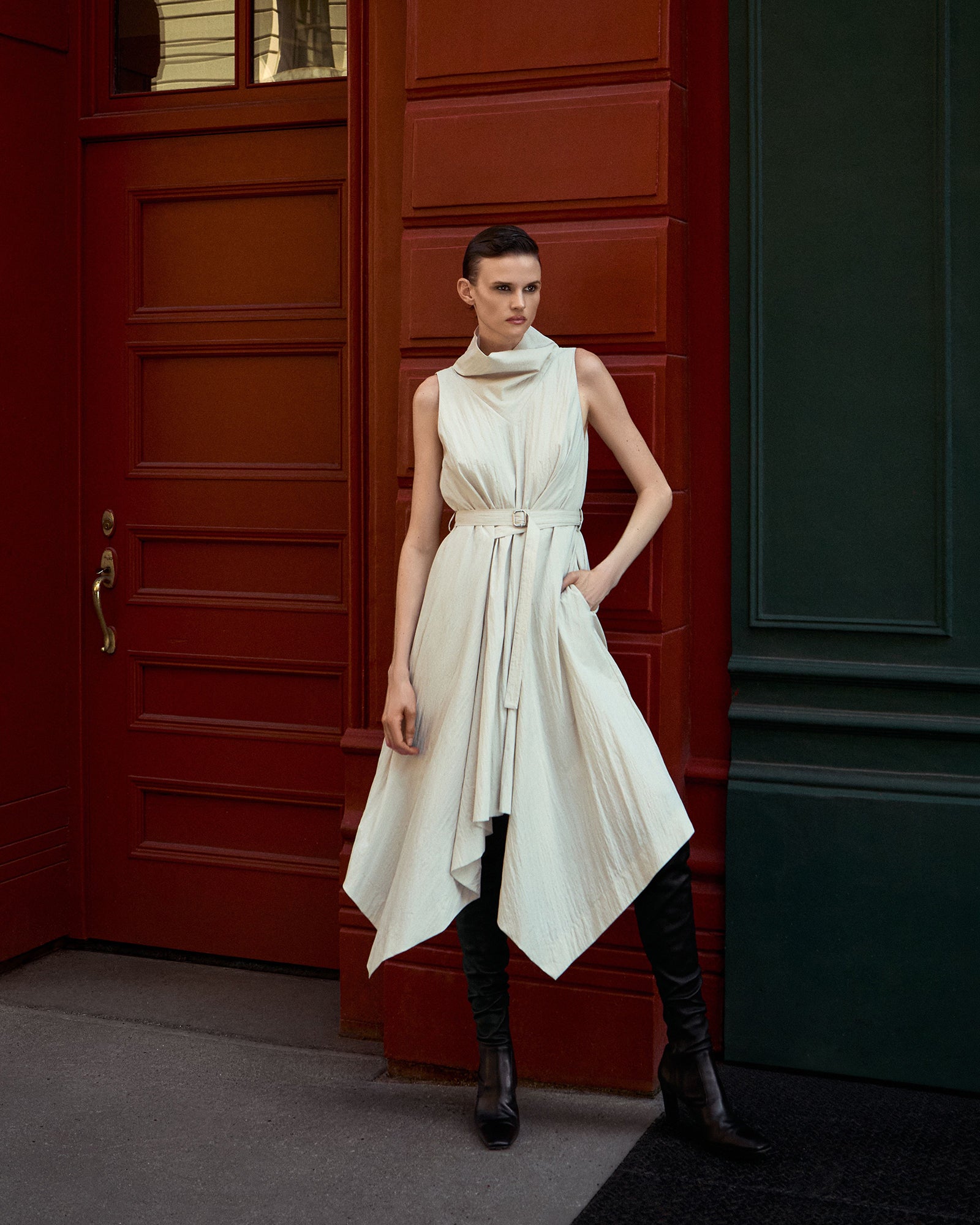 A model stands in front of a red paneled door wearing a sleeveless, high-neck, belted off-white dress with an asymmetrical draped hem, paired with black knee-high boots.