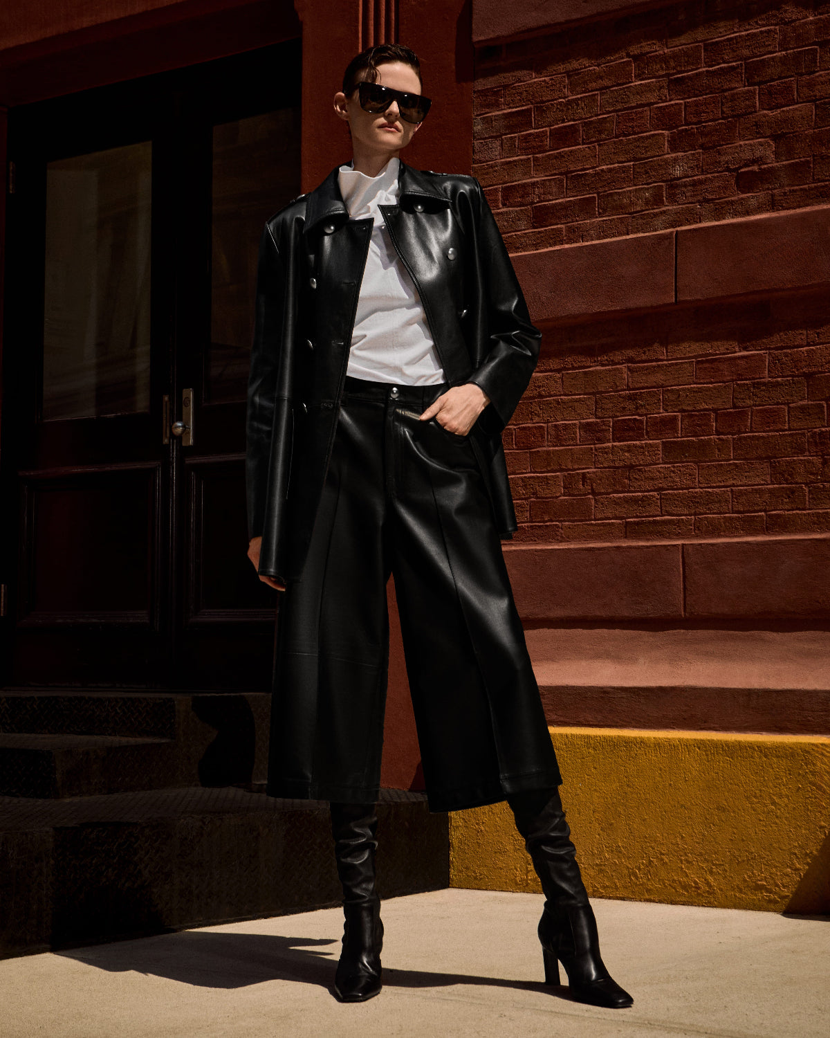 A model wearing a black leather jacket, matching wide-leg leather pants, a white shirt, and black knee-high boots, standing on outdoor steps in front of a brick building.