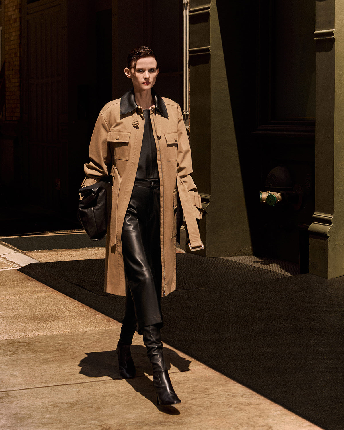 A model walks down a sunlit city street wearing a long tan trench coat over a black outfit, with black leather pants and tall black boots, and carrying a black shoulder bag.
