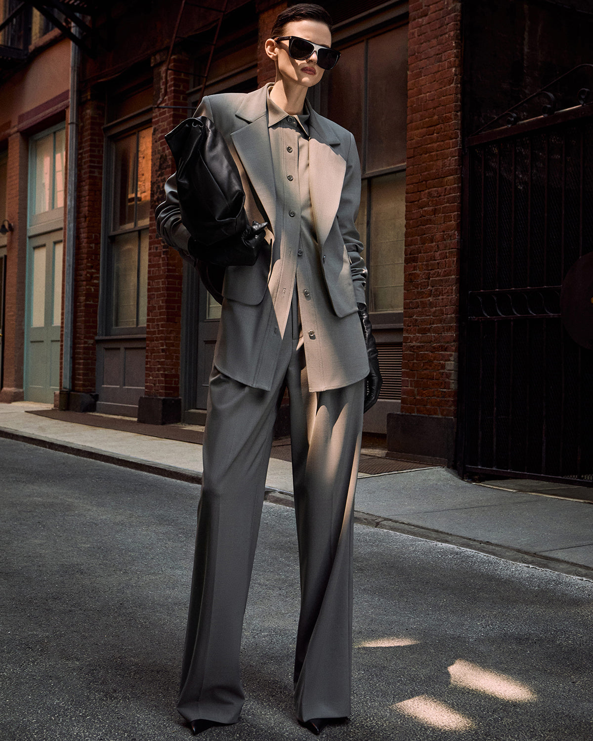 A model wearing a modern, tailored gray suit with wide-leg trousers, a structured blazer, and a matching vest, accessorized with a black leather shoulder bag and sunglasses, standing on an urban street.
