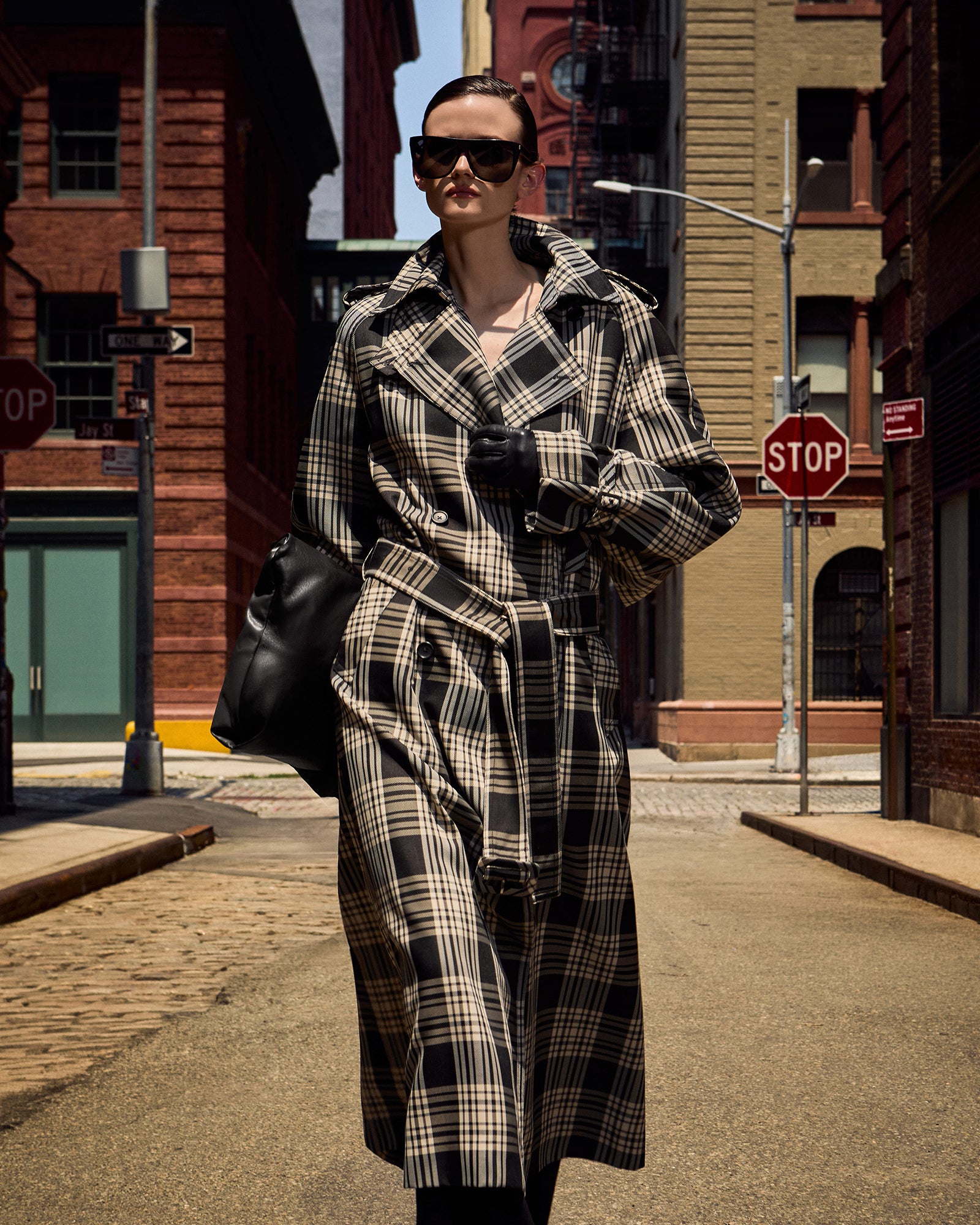 A woman walking down a city street wearing a long black and beige plaid trench coat, black sunglasses, and carrying a black shoulder bag.