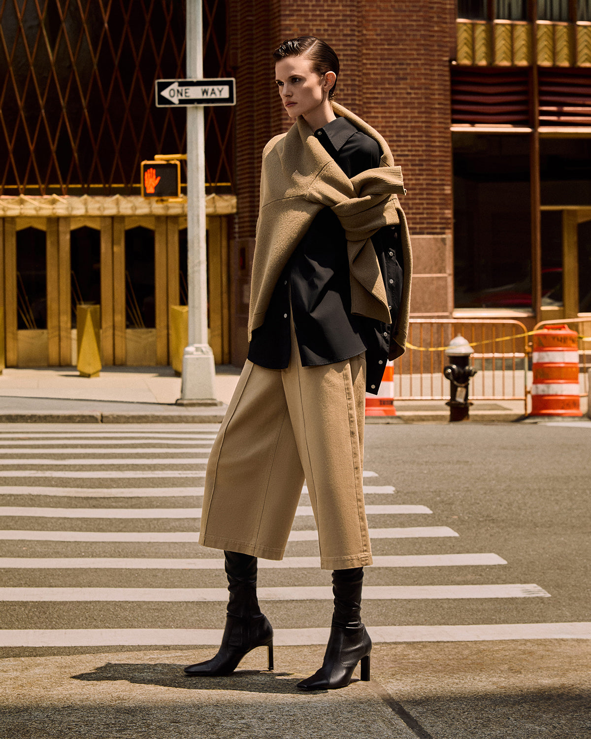 A model stands on a city crosswalk wearing a beige wrap coat over a black blazer, wide-leg beige pants, and black heeled ankle boots, with a city street and brick building in the background.