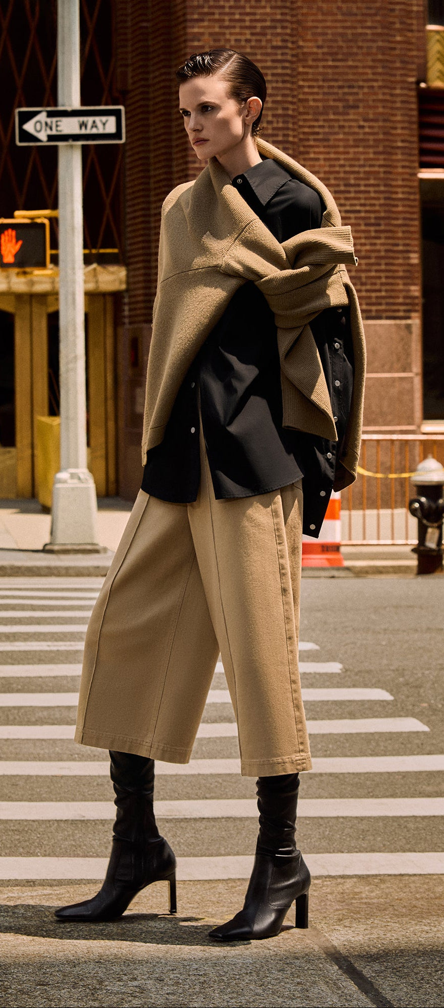 A model stands on a city crosswalk wearing a beige wrap coat over a black blazer, wide-leg beige pants, and black heeled ankle boots, with a city street and brick building in the background.