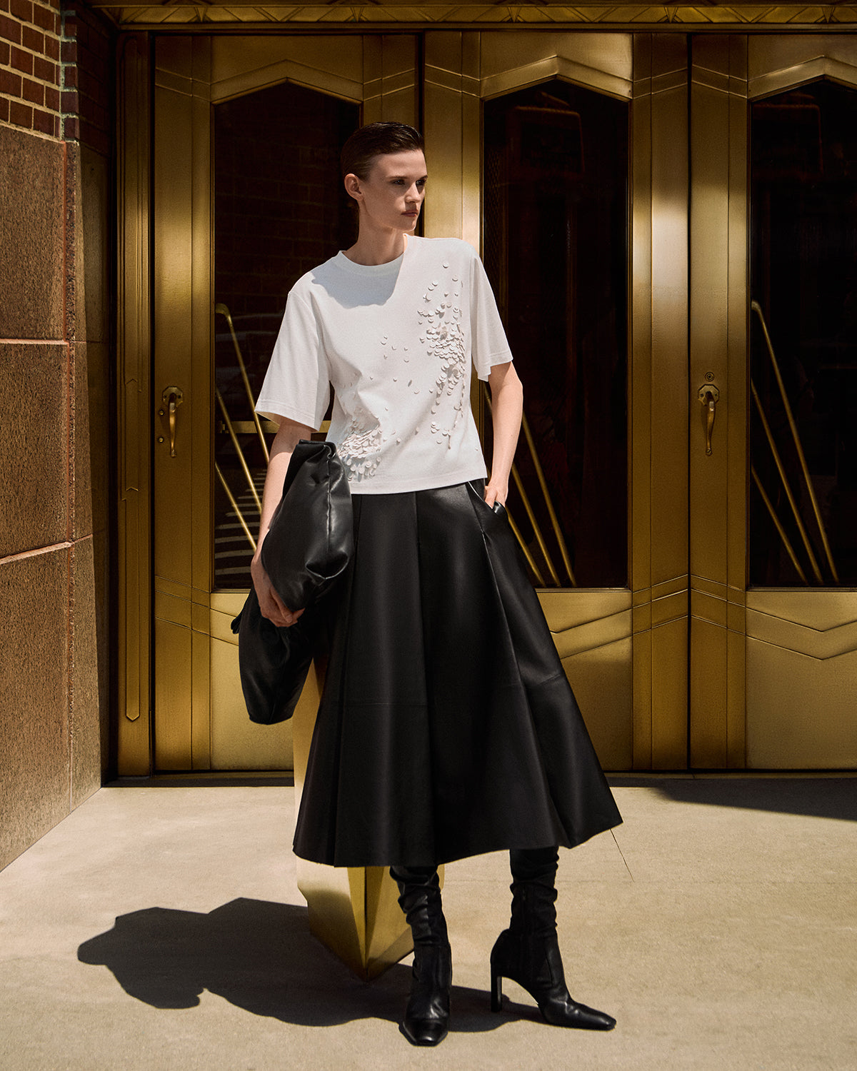 A model stands in front of gold-toned double doors wearing a white short-sleeve T-shirt with embellished detailing on the front, a black pleated midi skirt, and black knee-high heeled boots, while holding a black clutch.