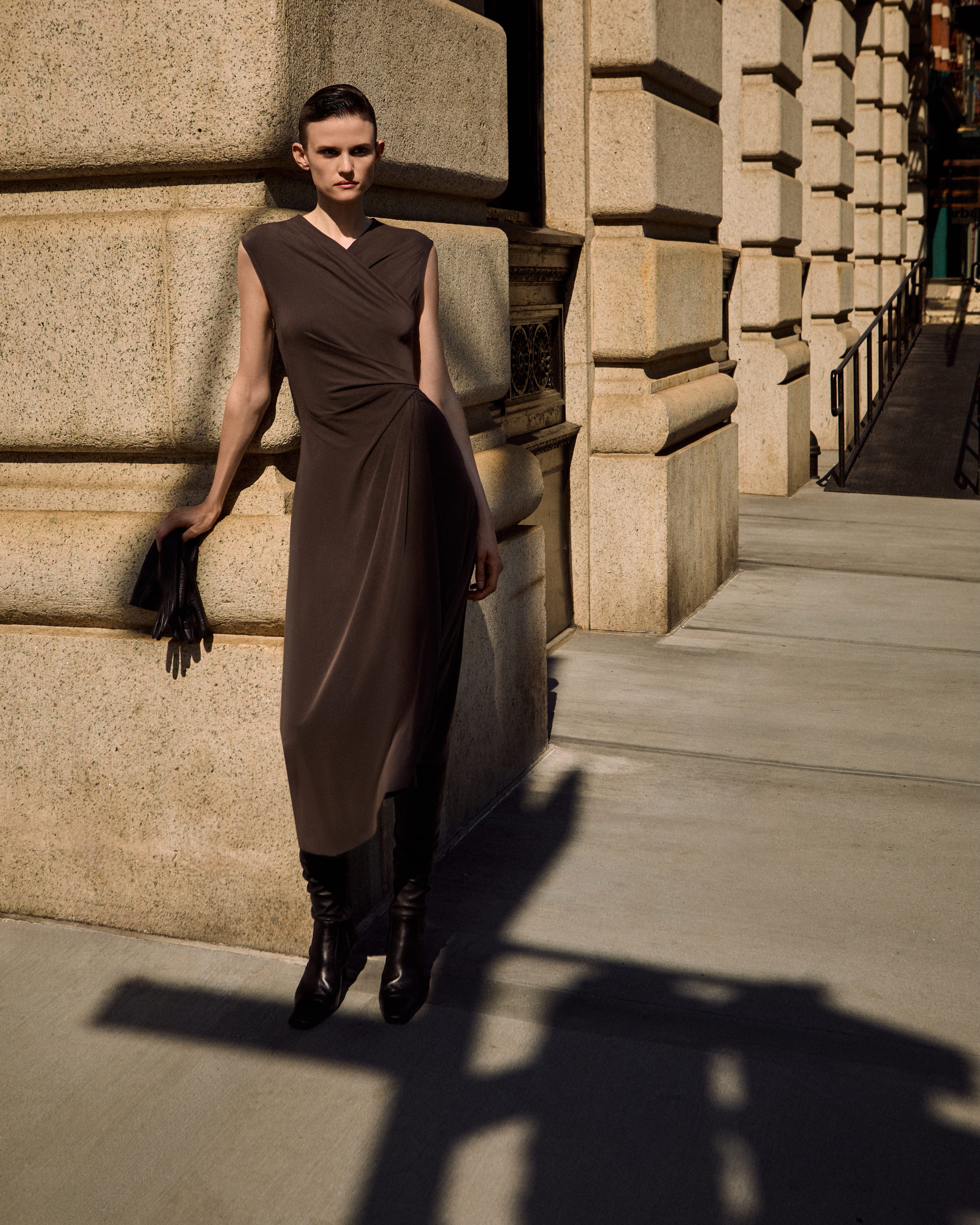 A model wearing a sleeveless, wrap-style brown dress and black ankle boots stands on a city sidewalk next to a stone building, holding a black clutch.