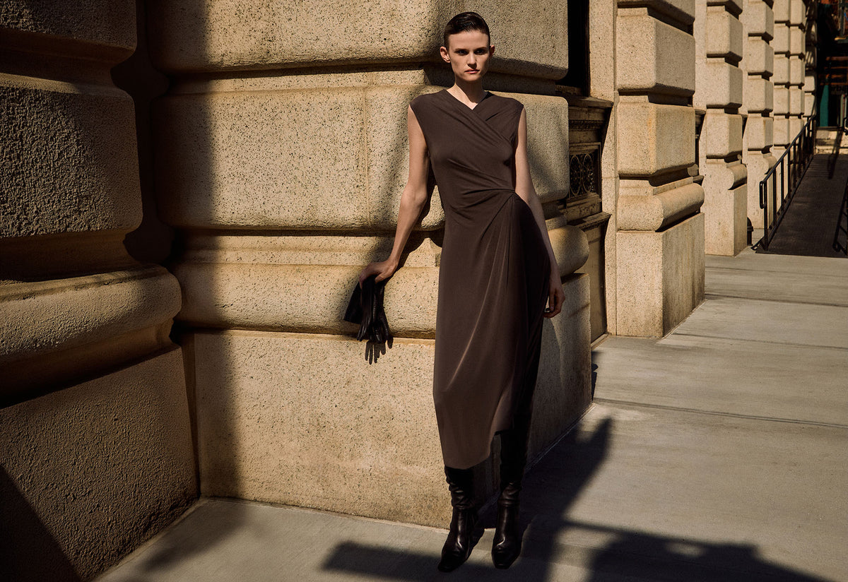 Model wearing a draped brown dress with brown gloves standing outside next to a building.
