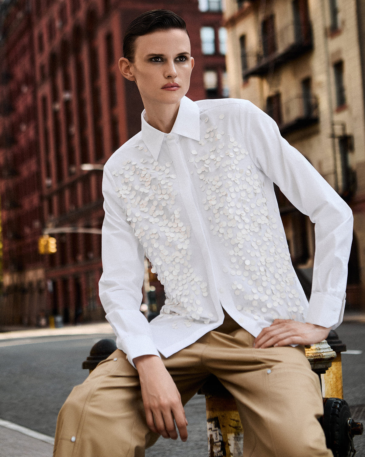 A model wearing a white long-sleeve button-up shirt with intricate 3D floral applique on the front, paired with beige cargo pants, sitting on a street in an urban setting.