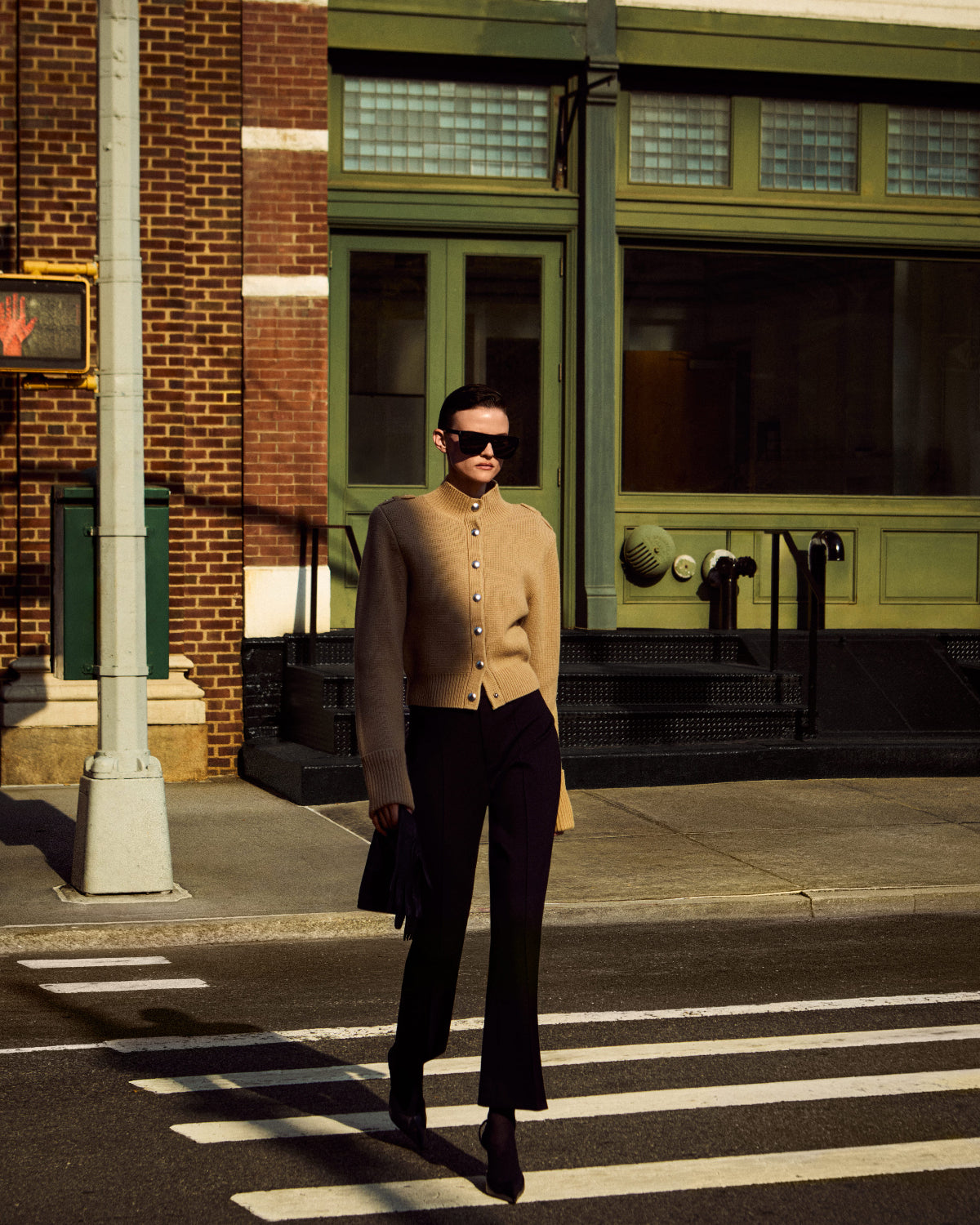 A person walking across a city crosswalk wearing a beige mock-neck button-front cardigan, black flared pants, black boots, and black sunglasses, with a brick building and green storefront in the background.