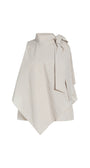 An asymmetrical cream-colored cape top with a high mock neck, textured diagonal crinkle pattern, and a large side bow detail, shown on a plain white background.